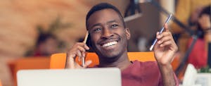 Young man speaking on the phone while sitting in front of a laptop and considering what to do when you can't get a cosigner.