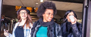 Three young women exiting an airport or train station, cellphones or coffee in hand