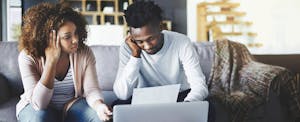 high-earners-struggle-emergency-costs-credit-karma A young high-earning couple anxiously discusses their finances on the couch. They weren't expecting so many unexpected expenses and emergency costs to come up in one year and they need to borrow money to pay for it all.