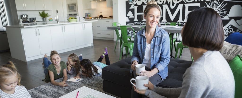 Two women having coffee, seated on sofas, while their daughters are coloring on the floor nearby