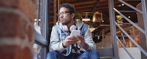 americans-savings-up Man sitting on steps, holding his phone and looking out the window, looking thoughtful