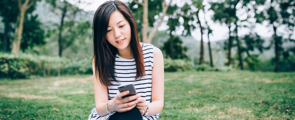 Young woman sitting outside on the grass, looking up how does a savings account work on her cellphone