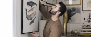 Man staging home carefully hangs a painting of whales alongside other artwork.
