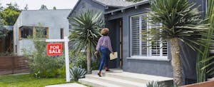 Woman carrying documents, including a seller's disclosure, walks into a house with a "for sale" sign on the lawn.