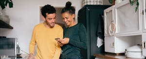 Two people standing together in their kitchen, smiling and looking at a cellphone together