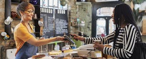 A woman smiles as she uses Apple Pay to purchase coffee from a barista at a cafe.