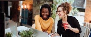 Two women sitting together at work, collaborating on their laptops
