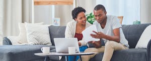 A couple seated on their living room sofa review their finances using a laptop and documents.