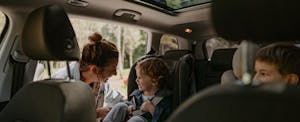 california-car-insurance-rates A mother buckles a small child into a carseat in the backseat of a car while another child looks on.