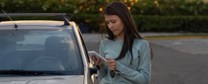 A woman checks standing next to her car uses a smartphone to check her car insurance rate.