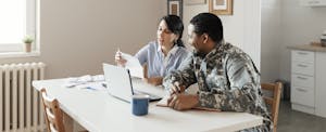 A man wearing military fatigues and his spouse sit at their dining room table and use a laptop to look into personal loan options.