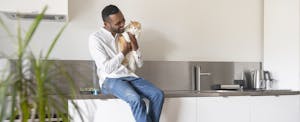Portrait of smiling man sitting on kitchen counter at home cuddling his cat