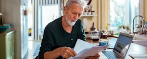 Older man in his kitchen, reviewing is bills at this kitchen counter, considering a debt settlement letter