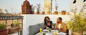 how-to-save-on-travel-despite-high-costs Couple smiling at each other as they sit together at a rooftop restaurant while on vacation