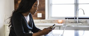 A person seated at a kitchen table holds their mobile phone while using a laptop to read about fiat currency.